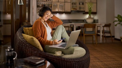 Femme assise dans son canapé face à son PC, téléphone en main